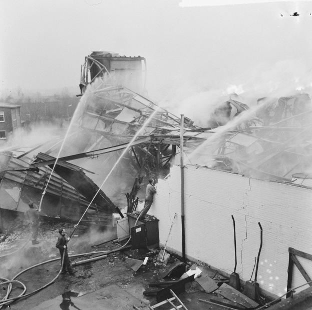 Schwarz-weiß-Foto von Feuerwehrleuten auf einem Dach, die ein Feuer löschen, während Menschen unten mit Leitern und Rohren helfen.