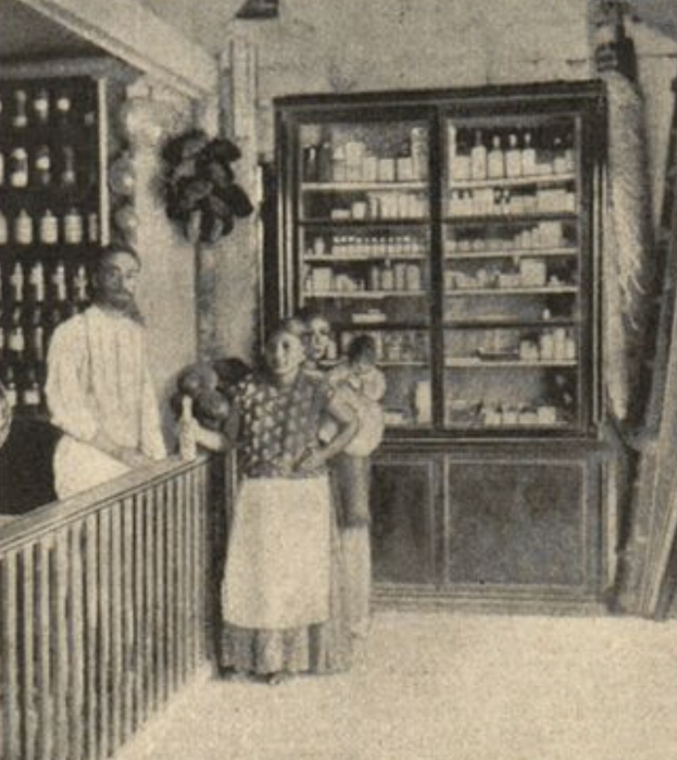 Schwarz-weiß-Fotografie einer Familie vor einer Apotheke mit einem Geländer auf der linken Seite und Regalen mit Flaschen im Hintergrund sowie Text am unteren Rand.