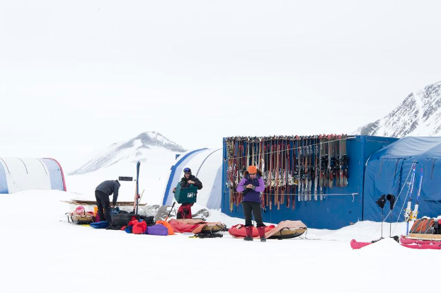 Drei Personen stehen auf einem schneebedeckten Eisfeld mit verstreuten Taschen, im Hintergrund Zelte mit Skiern darauf und schneebedeckte Hügel unter einem klaren Himmel.