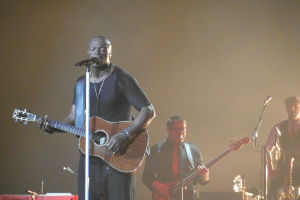 Ein Mann steht auf der Bühne und spielt eine Gitarre, während er in ein Mikrofon singt und von anderen Musikern umgeben ist, die verschiedene Instrumente spielen, einschließlich eines Saxofons.