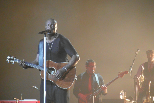 Ein Mann steht auf der Bühne und spielt eine Gitarre, während er in ein Mikrofon singt und von anderen Musikern umgeben ist, die verschiedene Instrumente spielen, einschließlich eines Saxofons.