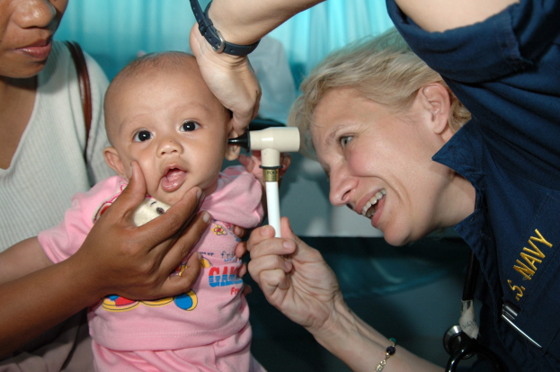 Eine Frau hält ein Baby, während ein Arzt es mit einem Otoskop untersucht, wobei eine Frau im Hintergrund mit einem Stethoskop assistiert.