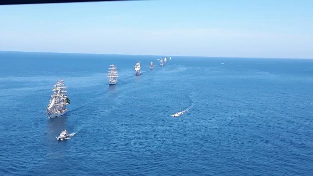 Eine große Gruppe von Segelschiffen auf dem Meer während eines Regattas, mit einem klaren blauen Himmel und hellen Sonnenstrahlen, die auf dem Wasser reflektieren.