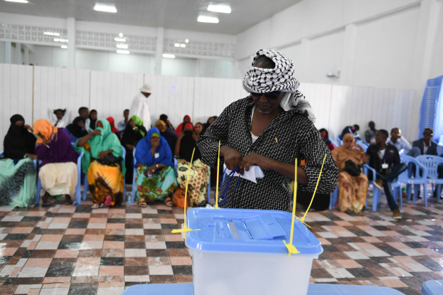 Eine Frau gibt ihre Stimme in einem Wahllokal in Mogadischu, Somalia ab, vor einem Tisch mit einer Box darauf sitzend, umgeben von Personen, mit einer Person im Hintergrund und von Deckenlampen beleuchtet.