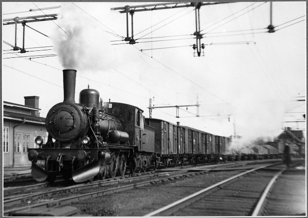 Schwarz-weiß-Foto eines Dampfzugs auf Schienen mit Rauch aus seinem Schornstein, ein Gebäude auf der linken Seite und Strommasten mit Drähten im Hintergrund.