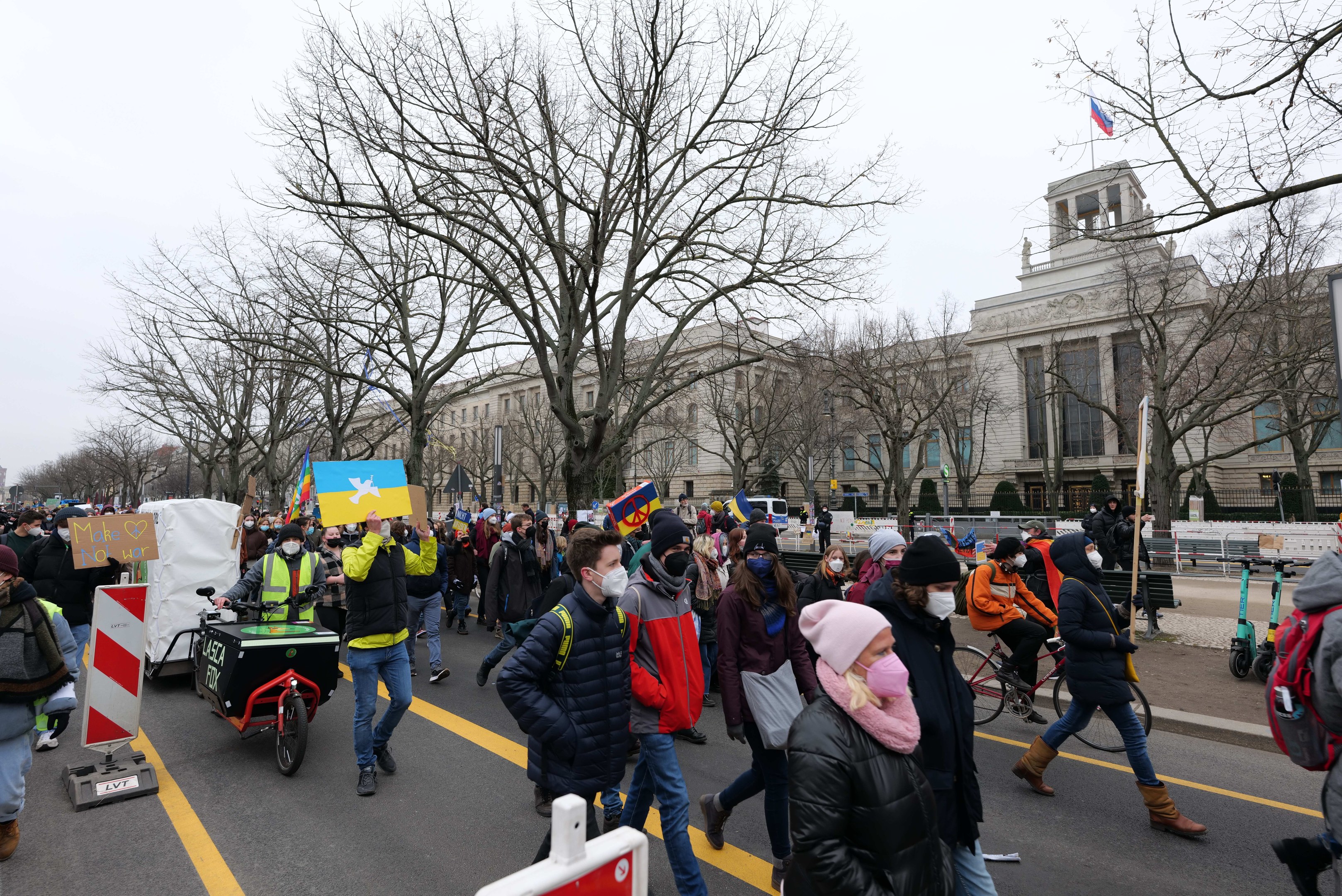 Eine große Protestmarsch mit Menschen, die eine Straße in Washington, D.C. entlanggehen, einige halten Schilder und andere fahren Fahrräder, mit Bäumen und einem klaren blauen Himmel im Hintergrund.