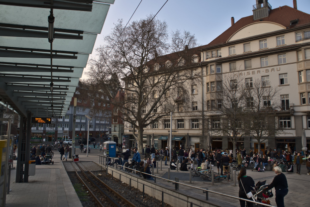 Eine belebte Straßenkreuzung mit einem Bahnhof auf der linken Seite, umgeben von Gebäuden, Bäumen, Laternen und Texttafeln, wo Menschen gehen, auf Bänken sitzen oder stehen, unter einem sichtbaren Himmel.