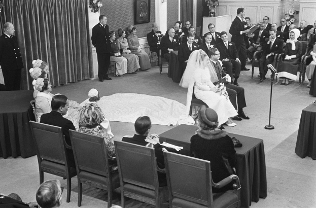 Schwarz-weiß-Foto einer Hochzeitszeremonie mit Menschen, die sitzen und stehen, einem Mann mit Mikrofon, einer Frau mit Blumenstrauß, einem Podium mit Mikrofon, Blumenschmuck, einem Vorhang, einem Rahmen an der Wand und einer Tür im Hintergrund.