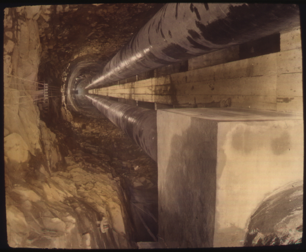Ein Schwarz-Weiß-Foto eines Tunnels mit einem großen Rohr, das hindurchläuft und von Felsen auf der linken Seite umgeben ist.
