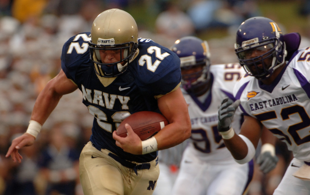 Ein Fußballspieler in einem marineblauen Trikot und Helm rennt mit dem Ball während eines Spiels, mit einem unscharfen Hintergrund, der schnelle Bewegung andeutet.