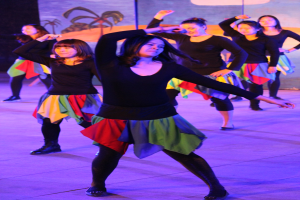 Junge Frauen in bunten Röcken und Schuhen tanzen energisch auf einer Bühne mit einem sichtbaren Gemälde im Hintergrund.