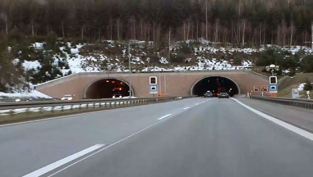 Autobahn mit Autos, die durch einen von Geländern und Schildern gesäumten Tunnel fahren, Schnee auf dem Boden, Bäume und Masten im Hintergrund und Himmel darüber sichtbar.
