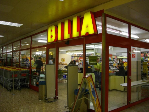 Ein Markt mit einem Eingang in der Mitte, Mülltonnen daneben, einer Reihe von Einkaufswagen links und ein paar Menschen drinnen, die unter Deckenbeleuchtung Produkte anschauen.