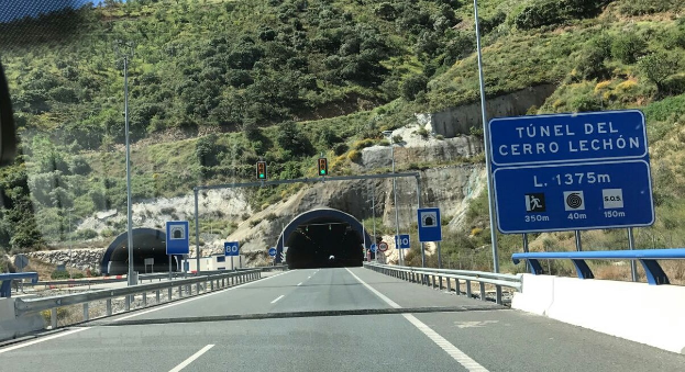 Ein Auto fährt durch einen Tunnel auf einer Straße mit Geländern, Schildern, Pfosten, Verkehrszeichen, Bäumen, Felsen und einem klaren blauen Himmel.