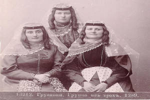 Drei Frauen in traditionellen russischen Hochzeitskleidern mit aufwendiger Stickerei und Perlenposen sich vor einer weißen Wand in einem Schwarz-Weiß-Foto.
