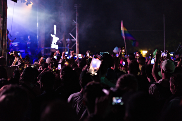 Menge an Menschen bei einem Konzert mit hochgehaltenen Handys, beleuchtet von Lichtern und umgeben von Pfählen und einer Fahne, während Musiker auf der Bühne spielen.