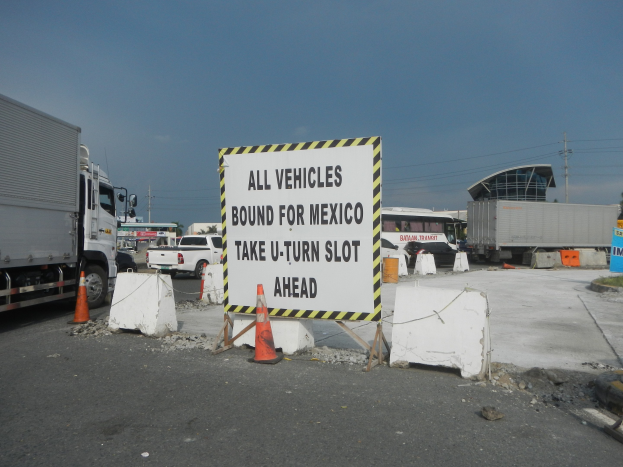 Lkw auf einem Parkplatz neben einem Schild mit der Aufschrift "Alle Fahrzeuge nach Mexiko" geparkt, mit verstreuten Verkehrskegeln, Brettern und anderen Gegenständen sowie im Hintergrund Gebäude, Bäume, Pfosten und einen klaren blauen Himmel.