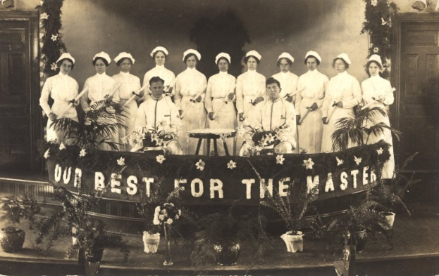 Eine Gruppe von Frauen in weißen Kleidern und Hauben steht auf einer Bühne mit einem Banner 'Unser Bestes für den Meister', umgeben von Blumentöpfen und mit Türen und einer Wand im Hintergrund.