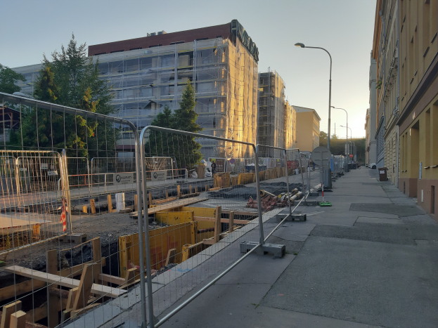 Baustelle in der Mitte einer Stadtstraße umgeben von Gebäuden, Fahrzeugen, Straßeninfrastruktur, Bäumen und einem klaren Himmel.