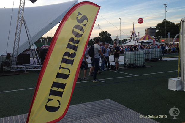 Ein lebhaftes Volksfest-Szenario mit Zelten, einem zentralen Mast, Menschen, Bäumen, Strommasten, Straßenbahnen auf der Straße und einem Eisengestell auf der linken Seite unter einem klaren Himmel.