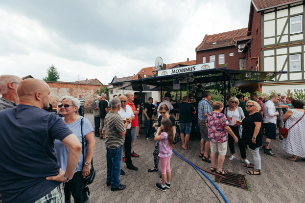 Gruppe von Menschen auf einem Bierfest vor einem Gebäude mit Fenstern, Bäumen und einem bewölkten Himmel, einige halten Gläser, mit einer Hütte mit Namensschild im Hintergrund.