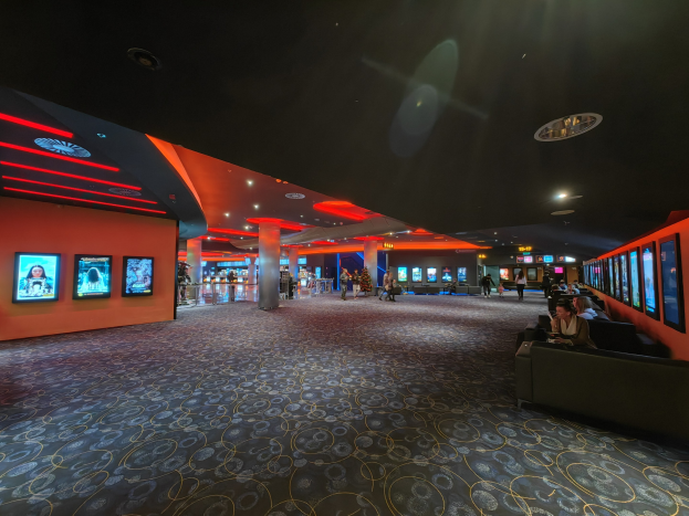 Ein geräumiger Raum mit Menschen auf Sofas sitzend, die Fernsehgeräte an den Wänden anschauen, Säulen verteilt und Deckenlichter, mit dem Boden unten sichtbar.