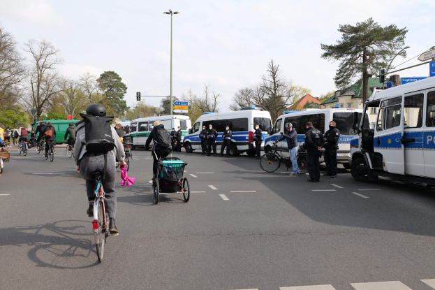 Eine Gruppe von Menschen, die auf Fahrrädern eine Straße entlangfahren, mit Polizeiwagen, einige tragen Helme und haben Taschen oder schieben Kinderwagen, mit Bäumen, Gebäuden und Verkehrszeichen im Hintergrund.