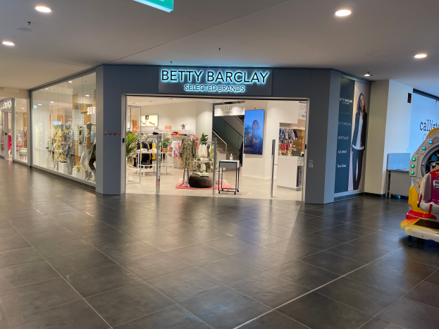 Das Innere eines Betty Barclay-Geschäfts in einem Einkaufszentrum mit Schaufensterpuppen in Kleidern, hängenden Kleidern, Tischen, Pflanzen und einem Spielzeugfahrzeug auf der rechten Seite, mit einer Tafel, die "Betty Barclay Selected Brands" anzeigt.