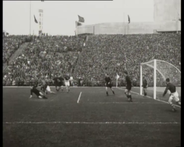Ein Schwarz-Weiß-Foto eines WM-Finale-Soccer-Spiels 1958 zwischen Manchester United und Liverpool mit Spielern auf dem Feld, einem Torpfosten auf der rechten Seite, Zuschauern auf den Tribünen und Fahnen im Hintergrund.
