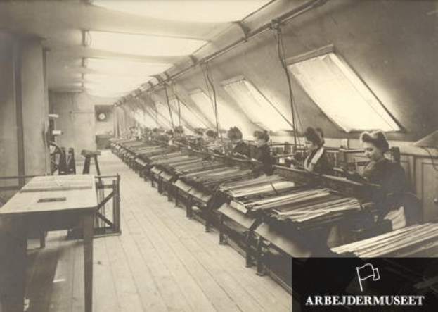 Ein Schwarz-Weiß-Foto von Frauen, die in einer Textilfabrik arbeiten, die an Maschinen sitzen, mit Tischen auf der linken Seite, einer Uhr an der Wand im Hintergrund und Text in der rechten unteren Ecke.