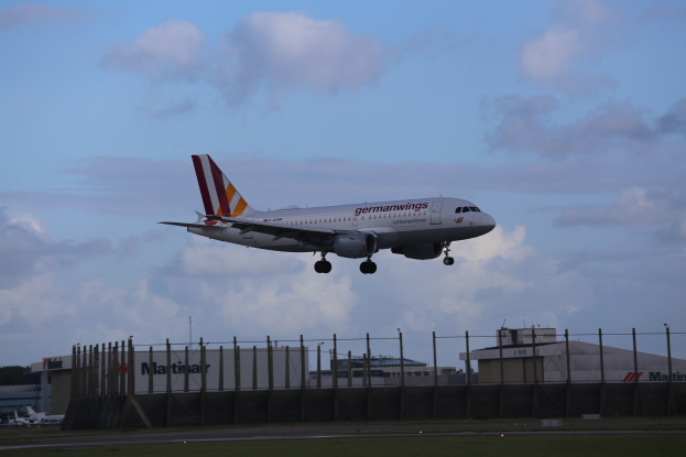 Germanwings Airbus A320-200 beim Start am Frankfurter Airport mit Gras darunter, einem Zaun im Vordergrund, Gebäuden und Bäumen im Hintergrund und einer bewölkten Himmel überhaupt.