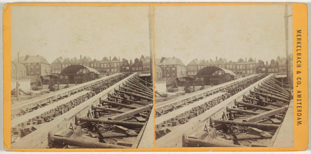 Ein altes Schwarz-Weiß-Foto einer Bahnschiene mit Gebäuden im Hintergrund, Text auf der rechten Seite mit der Aufschrift "Merkelbach & Co, Co. Amsterdam" und verstreuten Holzobjekten im Vordergrund.