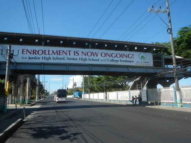 Ein Bus fährt unter einer Brücke mit einem Schild, auf dem "Anmeldung jetzt offen" steht, während Füßgänger darunter gehen und Strommäste an der Straße entlanglaufen, mit Bäumen und einem klaren Himmel im Hintergrund.