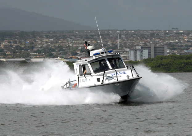 Polizeiboot fährt durch Wasser in städtischer Umgebung mit Passagieren, umgeben von Bäumen, Gebäuden, Strommasten, Bergen und einem klaren Himmel.