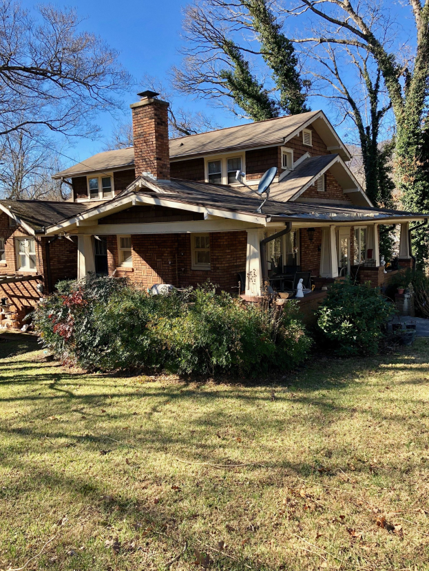 Ein Haus mit einem durch einen Sturm beschädigten Dach, umgeben von Pflanzen, Gras, Bäumen und unter einem bewölkten Himmel.