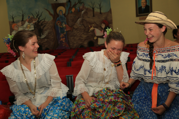 Drei Frauen in weißen Oberteilen und Röcken sitzen und lachen, mit roten Stühlen und Kunst an der Wand im Hintergrund.