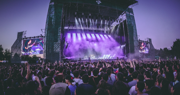 Eine große Menge schaut einer Band zu, die auf einer hell erleuchteten Bühne an einem Musikfestival spielt, mit Bäumen, einem Gebäude und einem klaren blauen Himmel im Hintergrund.