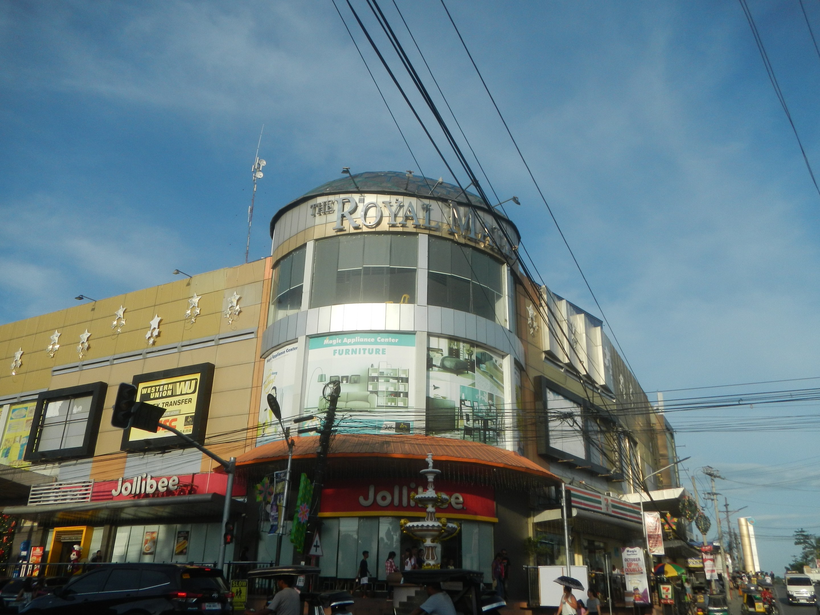 Eine belebte Stadtstraße mit fahrenden Fahrzeugen, Fußgängern auf dem Gehweg und einem großen Gebäude mit der Aufschrift "Royal Mall" im Hintergrund, umgeben von parkenden Autos, Bäumen, Strommasten und Drähten unter einem klaren Himmel.
