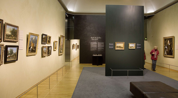 Großer Raum mit zahlreichen Gemälden an den Wänden, zwei Personen, eine Couch, ein Tisch mit Gegenständen, eine Tafel mit der Aufschrift "Karel du Jardin Ausstellung in der Nationalgalerie der Künste" und Deckenbeleuchtung.