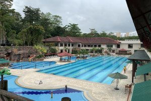 Großes Schwimmbad im Ritz-Carlton, Kota Kinabalu, mit Menschen, Sonnenschirmen, Stühlen, Tischen, Pflanzen, Bäumen, Felsen, Gebäuden, einem Schuppen auf der rechten Seite und Himmel im Hintergrund.