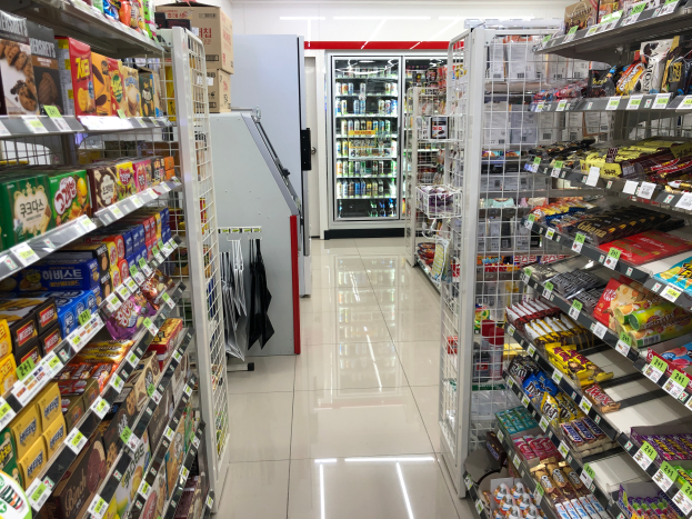 Ein Supermarktgang mit verschiedenen Lebensmittelarten auf Regalen mit Preisschildern, ein Kühlschrank im Hintergrund und Deckenleuchten.