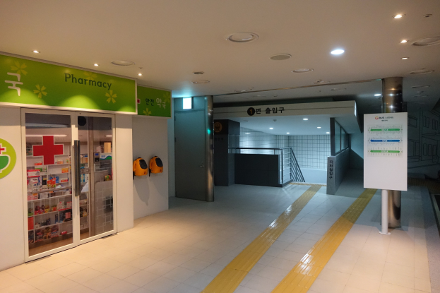 Apothekeneingang mit einem "Apotheke"-Schild links, einer Tafel mit Text rechts, beleuchteter Dachbeleuchtung und Hintergrundobjekten.