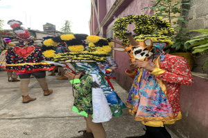 Eine Gruppe von Menschen in bunten Kostümen steht vor einem Gebäude während eines Faschingsumzugs, mit verschiedenen Gegenständen, Bäumen, Gebäuden und einem klaren blauen Himmel im Hintergrund.