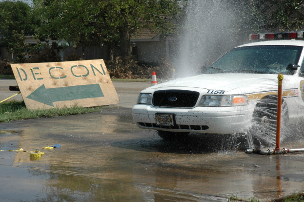 Polizeiauto fährt durch eine Pfütze mit verstreuten Trümmern, darunter ein Verkehrskegel und eine Tafel mit Text, vor dem Hintergrund von Gebäuden, Bäumen und einem klaren Himmel.