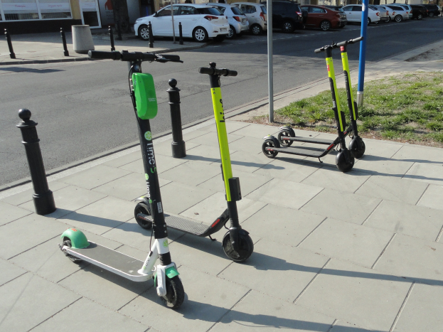 Drei Elektro-Scooter auf einem grasbewachsenen Straßenrand mit Fahrzeugen, Gebäuden, Bäumen und einem klaren Himmel im Hintergrund.
