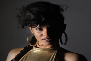 Eine Frau in einem goldenen Kleid und großen Kreolen steht selbstbewusst vor einer Wand, ihre Haare sind zu einem glatten Bob gestylt und sie schaut direkt in die Kamera mit einem starken und bestimmten Ausdruck.