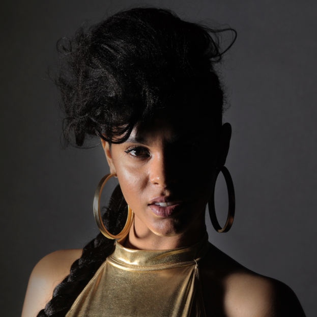 Eine Frau in einem goldenen Kleid und großen Kreolen steht selbstbewusst vor einer Wand, ihre Haare sind zu einem glatten Bob gestylt und sie schaut direkt in die Kamera mit einem starken und bestimmten Ausdruck.