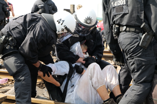 Eine Gruppe von Polizisten in Einsatzausrüstung verhaftet einen Mann in einem weißen Kleid auf einem Bahnsteig, mit anderen im Hintergrund sichtbar.