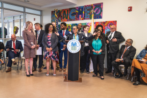 Gruppe von Menschen vor einem Podium mit Mikrofon und einer Tafel mit dem Text "Sancho-Stadtrat tritt dem Stadtrat bei", mit sitzenden und stehenden Teilnehmern, Flaggen, Feuerlöscher und Deckenbeleuchtung.