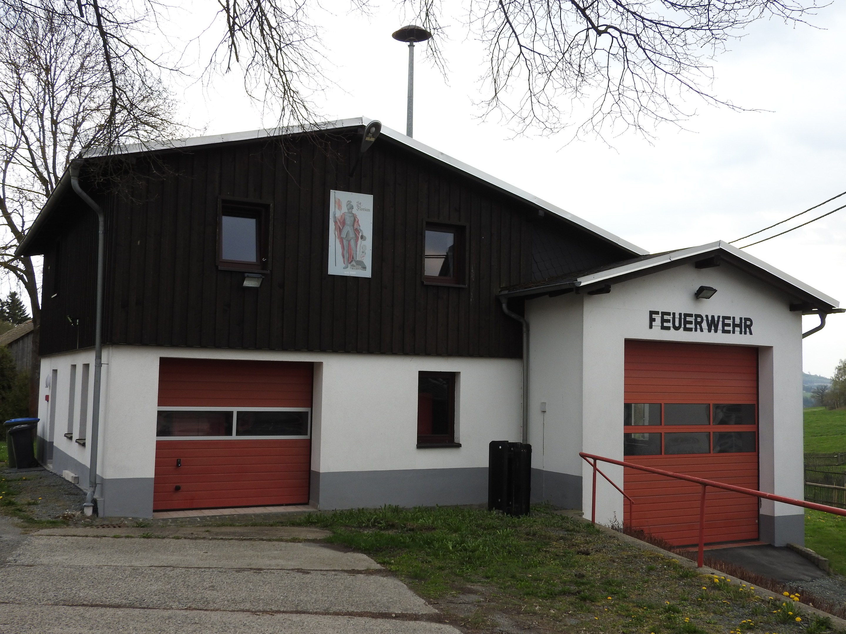 Feuerwache mit rotem und weißem Gebäude, Straße, Gras, gelben Blumen, schwarzen Objekten, Geländer, Pfahl, Drähte, Bäume und Himmel im Hintergrund.